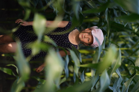 A little girl in a pink hat runs among the corn stalks across the field. She's wearing a black dress, she's smiling and looking at the camera, she's having fun. Knee-deep view. The leaves green partially block the view.の写真素材