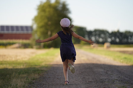 A girl in a blue dress and a pink hat runs along a dusty field road happily spreading her arms to the sides. Around the sunny day, trees and in the distance you can see the building. View of the running child from behind. fun holidays.の写真素材