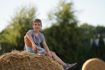 A teenager sits on a haystack on a sunny summer day and smiles at the camera. The guy sits with one leg crossed and his arm hanging down. In the background are haystacks and green trees. Fun childhood and holidays in the village.の写真素材