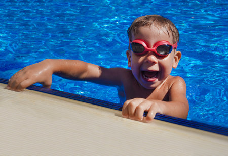 Happy boy with red goggles in open air swimming pool. Portrait of young boy leaning on edge of swimming pool on summer vacationの写真素材