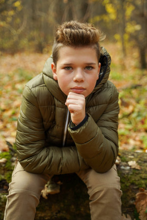 A cute stylish guy sits in the autumn forest propping up his chin with his hand and looks into the camera. Autumn fashion for men.の写真素材