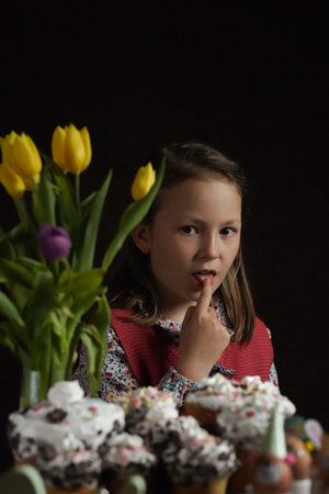 An attractive girl in a rustic outfit sits in front of a table with Easter decorations and licks the...