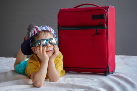 A little boy in a summer cap lies next to a suitcase and dreams of a summer vacationの写真素材