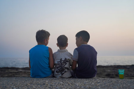Three boys sitting on the shore of the sunset sea, rear view.の写真素材