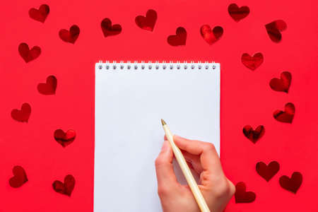 White blank notepad with woman hand holding pen on red background with red shiny hearts confettiの写真素材