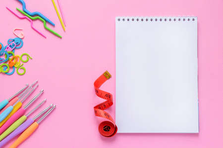 Top view of white empty notebook with crocheting and knitting accessories on soft pink background