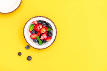 Directly above view of coconut bowl with fresh ripe berries on bright yellow background with copy...