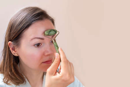 Young caucasian woman doing face massage with green jade roller on neutral background