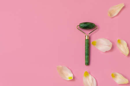 Facial roller made of natural green jade stone on pink background with white petals and copy spaceの写真素材