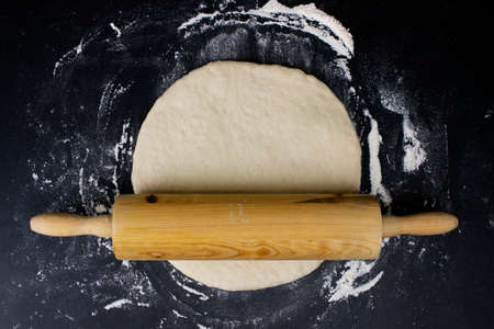 Rolling pin with dough and flour on black table. Top view