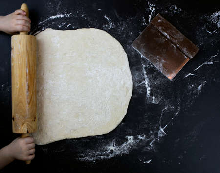 Rolling pin in kids hands with dough and flour on black table. Top view