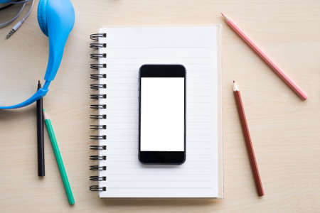 top view blank screen of mobile phone on notebook and wooden desk with pencil and headphone
