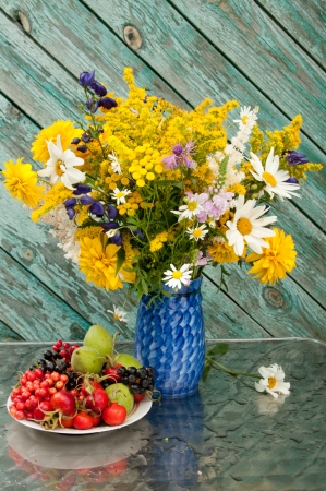 still life bouquet of chamomiles, yellow and blue flowers on the blue vase with berries on the plateの写真素材