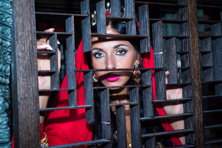 eyes beautiful Arab woman in red, peeping from behind a wooden lattice ornamentalの写真素材