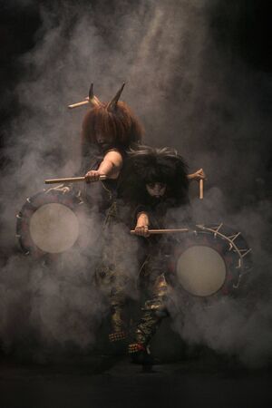 Two taiko drummers in a wig and a demon mask on stage with drumsticks and drums on a black background with smoke in light rays. Demons from Japanese mythology.の写真素材