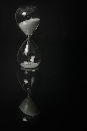 white sand hourglass on a black mirror surface with reflections isolated on black background.