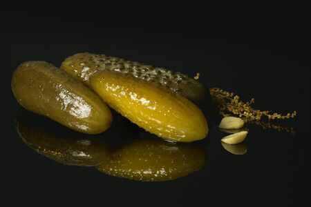 Three pickled cucumbers with garlic and estragon on a black mirror table with reflections isolated...