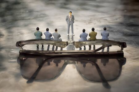 Miniature model of businessmen sitting on glasses backside and a presenter standing in front of them...