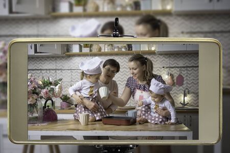 Young female blogger with grandmother and two daughters, live streaming a cooking with children show...