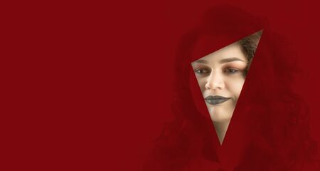 the face of a young beautiful girl with a bright make-up and with plump dark lips peeks into a hole in red paper. Fashion, beauty, make-up, cosmetics, beauty salon, style, personal care, geometry.の写真素材