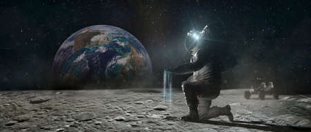 Astronaut sits on his knee on the lunar surface and pours blue earth's soil through his fingers. Planet Earth rise at the horizon.の写真素材