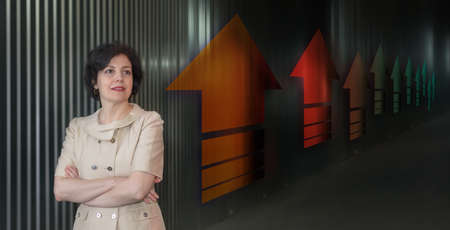 Pretty middle aged business woman in business suit, successful confidence close up head and shoulders portrait against dark wall with up arrows in perspective.の写真素材