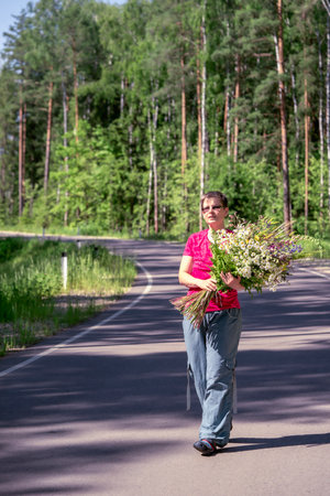 full-length portrait of a happy smiling woman with a huge bouquet of wild flowers walking along a forest roadの写真素材