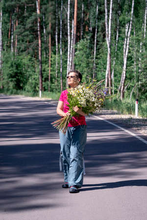 full-length portrait of a happy smiling woman with a huge bouquet of wild flowers walking along a forest roadの写真素材