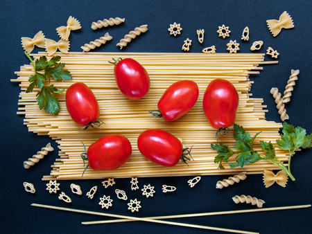 Spaghetti with tomatoes, greens and other figured pasta in the form of bows, spirals, figures of rockets and stars for cooking on a dark backgroundの写真素材