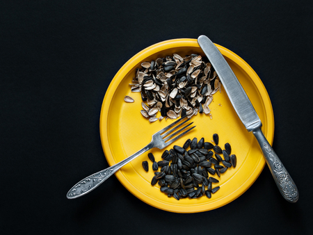 On a dark background there is a yellow plate with uncooked sunflower seeds, the husks from the seeds, a knife, a fork shot from above with a close-upの写真素材