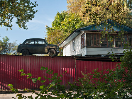 Near a white building with large windows behind a red fence on the roof of a shed stands an old retro car in the fall on a sunny day near trees with yellow leavesの写真素材