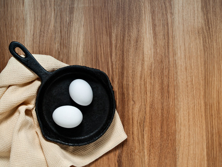 A black cast-iron pan with chicken eggs stands on a beige fabric napkin on a wooden background horizontally with copy spaceの写真素材
