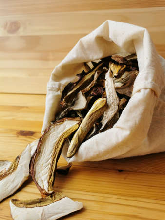 Close-up of dried chopped caps of porcini mushrooms and boletus mushrooms on a wooden table on a light wooden background from an open tucked linen bag.の写真素材