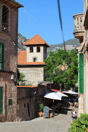 A narrow street in the town of Valldemossa with cafes, stone walls of houses and Windows with green...