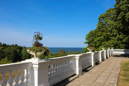 An outdoor terrace with a white curly fence, trees and a flowerpot against a bright blue sky. View...