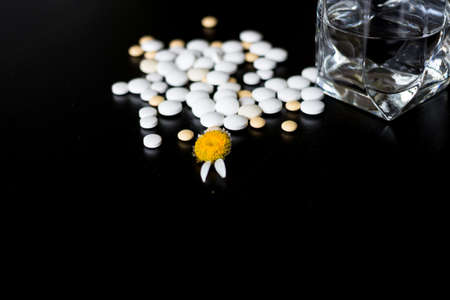 Chamomile with the remaining two petals. Next to it were pills of various sizes, taken from blisters and scattered on a black table, and a glass of water.の写真素材