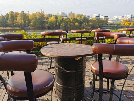 A deserted street cafe with high chairs and tables made of metal barrels on the riverbank in the...