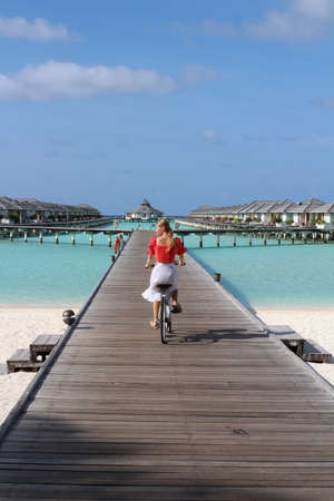 A blonde girl in a white skirt and a red blouse rides a bicycle in the afternoon along a gray wooden pier leading to villas above the Indian Ocean, standing on stilts, in the Maldives. Sun Island Resort and Spa Hotel.のeditorial素材