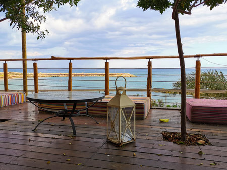 Protaras. Famagusta region. Cyprus. A cafe near the beach, without people, with soft, low mattresses, a decorative lantern, a glass round table and a tree growing inside against a blue sky with clouds.の写真素材