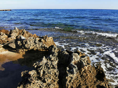 Protaras. Famagusta area. Cyprus. Long-hardened lava, porous, sharp coast of the Mediterranean Sea with incoming waves against a blue cloudless sky.の写真素材