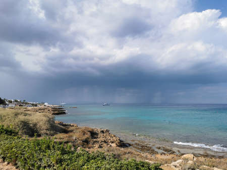 Protaras. Famagusta area. Cyprus. Sunny coast of the Mediterranean Sea with grass and stones, a dramatic sky and rain over the sea, a boat in the sea and the largest sailing yacht in the world.の写真素材