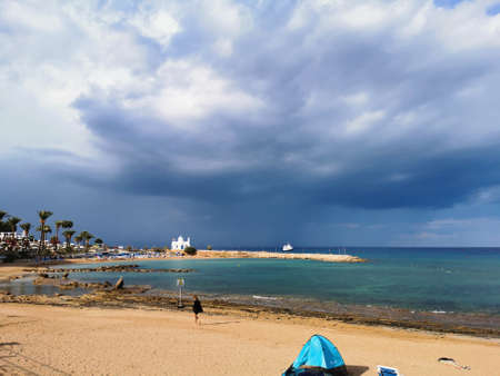 Protaras. Cyprus. The sunny sandy coast of the Mediterranean Sea, the beach in the bay, the temple of St. Nicholas the Wonderworker, a dramatic sky and rain over the sea, a ship in the sea.のeditorial素材