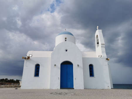 Protaras. Famagusta area. Cyprus. The Church of St. Nicholas the Wonderworker is white with a blue door against the backdrop of the Mediterranean Sea and the dramatic sky.の写真素材