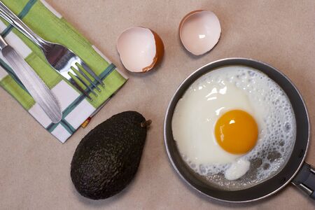 One fried egg on the little pan, fork and knife, avocado, egg shells, dish towel. Craft brown background. Copy space. Place for text and design. Top view. Flat lay.の写真素材