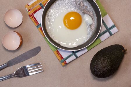 One fried egg on the little pan, fork and knife, avocado, egg shells, dish towel. Craft brown background. Copy space. Place for text and design. Top view. Flat lay.の写真素材