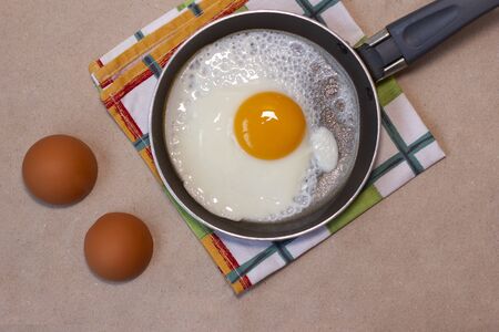One fried egg on the little pan, egg shells, dish towel. Craft brown background. Copy space. Place for text and design. Top view. Flat lay.の写真素材