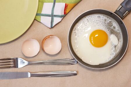 One fried egg on the little pan, fork and knife, green plate, egg shell, dish towel. Craft brown background.  Top view. Flat lay.の写真素材