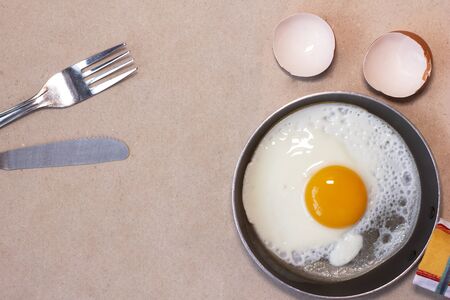 One fried egg on the little pan, fork and nife, egg shell, kitchen towel. Craft brown background. Copy space. Place for text and design. Top view. Flat lay.の写真素材