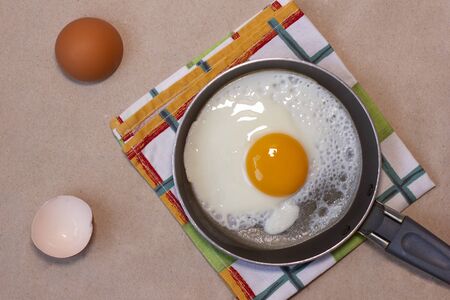 One fried egg on the little pan, dish towel, egg shells. Craft brown background. Copy space. Place for text and design. Top view. Flat lay.の写真素材