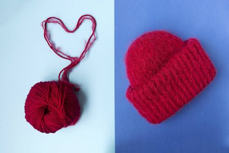 Knitted red hat on the two colors background. Blue background. Knitting courses. Online education. Flat lay. Top view. Place for text and design. Copy space. Knitting with love. Quarantine life. The heart of the threadの写真素材